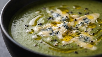 Superficie de una crema de brócoli verde con trozos de queso azul derretido, gotas de aceite de oliva y pimienta molida en un tazón.