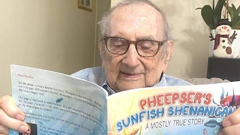 Primer plano de un anciano con gafas leyendo un libro infantil abierto con ilustraciones coloridas y texto. Al fondo, se ve un adorno de muñeco de nieve