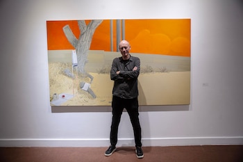 Hombre calvo con camisa oscura y pantalones negros de pie con los brazos cruzados frente a una pintura grande de un paisaje surrealista con cielo naranja