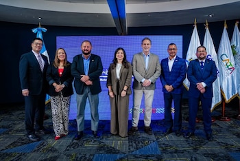 Siete personas de pie, seis hombres y una mujer. Detrás, una pantalla azul con texto y varias banderas blancas con los logos CATA y de Guatemala