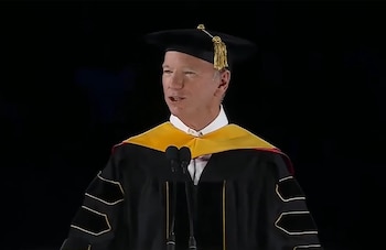 Eric Schmidt, con toga y birrete académico negro y estola amarilla, habla ante dos micrófonos sobre un fondo oscuro
