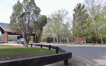 Entrada a un country con garita de seguridad y barreras. Un cartel de madera dice "Grand Bell Club de Campo". Hay árboles, pasto, camino y camioneta azul