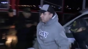 Un hombre con sudadera gris y gorra camina escoltado por agentes con uniforme oscuro en un entorno nocturno