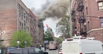 Imagen de un edificio de apartamentos del que emana humo negro espeso, con un camión de bomberos rojo con la escalera extendida en la calle