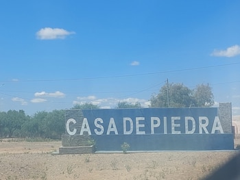 Un letrero de color azul con letras blancas que dicen CASA DE PIEDRA en un terreno árido con árboles y cielo despejado