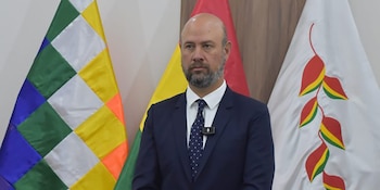 Hombre calvo con barba, traje oscuro, corbata y micrófono de solapa. Al fondo, bandera Wiphala, bandera boliviana y bandera blanca con diseño de hojas