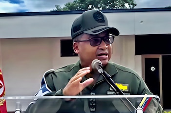 Un hombre con uniforme militar, gorra y anteojos habla frente a un micrófono en un podio transparente