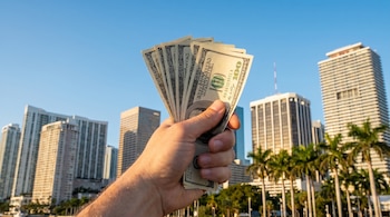 Una mano sostiene un fajo de billetes de cien dólares frente al skyline de Miami, con varios rascacielos y palmeras bajo un cielo azul claro.