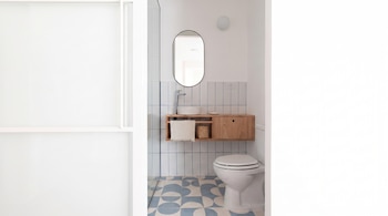 Vista de un baño con inodoro blanco, lavabo sobre mueble de madera, espejo ovalado y baldosas con patrones en azul y blanco.