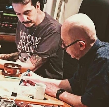 Primer plano de dos hombres en un estudio de grabación; uno calvo con gafas escribe, el otro con bigote y tatuajes lo observa desde atrás