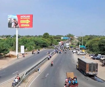Una carretera con multitudes de personas bloqueando el tráfico de vehículos, incluyendo camiones y coches parados. Un gran cartel publicitario rojo visible a la izquierda. Cielo azul