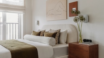 Cama con cabecero y ropa de cama blanca y verde oliva. Mesita de noche de madera con jarrón, flores, libros y reloj. Arte y lámparas de pared. Ventana con balcón.