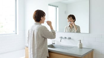 Mujer de cabello corto cepilla sus dientes frente a un espejo rectangular. En un baño blanco se observa un lavabo de cemento, un grifo y una ventana.
