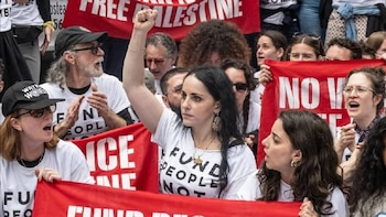 Una multitud de personas en una protesta, con una mujer en primer plano con el puño levantado. Se ven pancartas rojas con texto blanco, incluyendo "FREE PALESTINE"