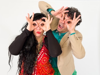 Un hombre y una mujer con las manos formando círculos alrededor de los ojos. Ella tiene pelo negro largo y labios rojos. Él viste un blazer beige y corbatín