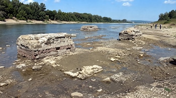 Tres estructuras de piedra sobresalen de un lecho de río expuesto con charcos de agua, con un río, árboles en el fondo y tres personas en la orilla.