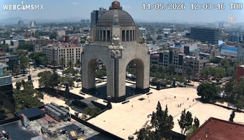 Vista aérea del Monumento a la Revolución, un gran arco de piedra con cúpula, rodeado de una plaza y edificios urbanos bajo un cielo despejado