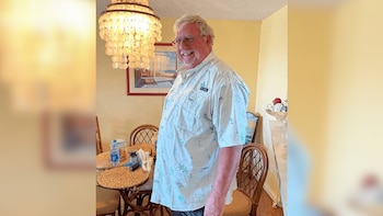 Un hombre caucásico de mediana edad con cabello blanco y gafas sonríe, vistiendo una camisa estampada, de pie en un comedor con un candelabro y una mesa de cristal