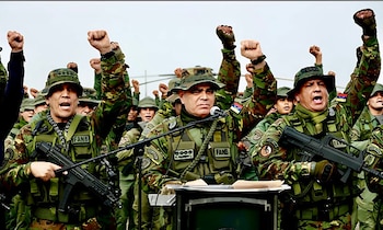 Varios militares con uniforme de camuflaje sostienen armas y levantan el puño cerrado ante un atril con micrófonos