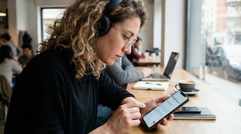 Primer plano de una mujer con cabello rizado, gafas y auriculares negros, usando un smartphone para una transcripción de audio a texto en un café.