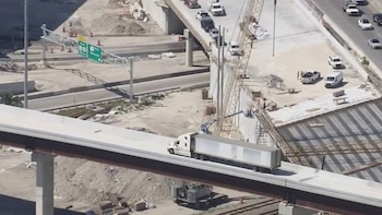 Vista aérea de un camión blanco con remolque gris circulando sobre un nuevo tramo elevado de autopista en construcción, con otras vías y obras en tierra