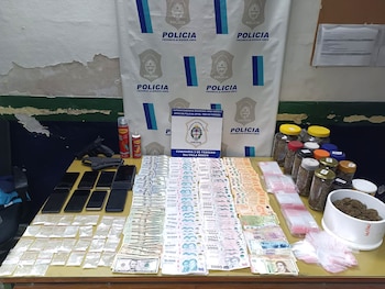 Mesa con billetes, teléfonos móviles, armas de fuego, bolsas pequeñas y frascos ante un cartel de la Policía de la Provincia de Buenos Aires