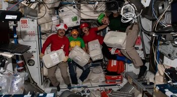Cuatro personas con disfraces y bigotes postizos flotan en el interior de una nave espacial junto a cables, aparatos y paquetes blancos