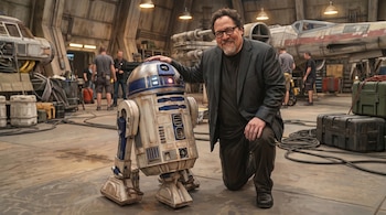 Ilustración de Jon Favreau arrodillado junto a R2-D2 en un hangar de Star Wars. Al fondo se ven técnicos y partes de naves espaciales.