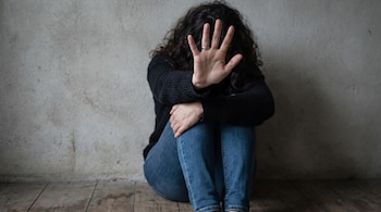 Mujer con cabello rizado oscuro, suéter negro y jeans azules, sentada en suelo de madera frente a pared. Mano derecha alzada con palma extendida.
