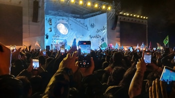 Vista desde la audiencia de un concierto nocturno, donde miles de manos levantan teléfonos móviles grabando un escenario iluminado con una gran huella dactilar