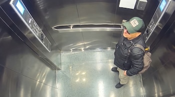 Vista superior de un hombre con gorra verde, chaqueta negra y mochila dentro de un ascensor metálico con paneles digitales.