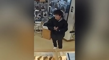 Una persona joven con cabello oscuro y chaqueta negra se para frente a estantes de mercancía y una vitrina con armas en una tienda