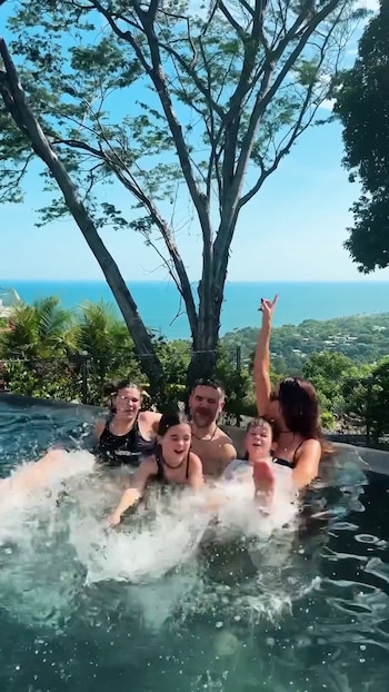 Cinco personas, dos adultos y tres niños, en una piscina con agua salpicando. Al fondo, vegetación y el océano bajo un cielo azul