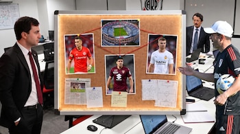 Tres hombres frente a un tablero de corcho con fotos de futbolistas, el estadio Monumental, diagramas de tácticas y notas manuscritas.