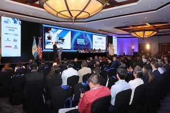 Hombre en podio, dos pantallas, panel de ponentes, banderas de Guatemala y Estados Unidos, auditorio con público sentado, candelabros
