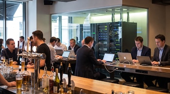 Personas con computadoras portátiles sentadas cerca de una barra de bar y una sala de cristal con bastidores de servidores.