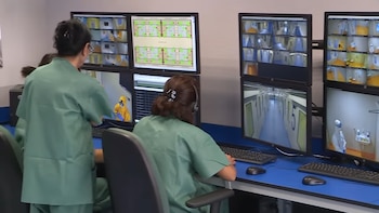 Dos personas en uniforme médico observan ocho monitores de seguridad que muestran imágenes de pasillos, habitaciones y personal en trajes protectores
