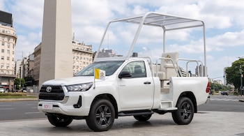 Camioneta blanca modificada con un sillón blanco, techo transparente y bandera papal junto al Obelisco de Buenos Aires.