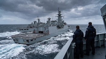 Dos buques militares navegan en el mar agitado bajo un cielo nublado, observados desde una cubierta por dos personas de espaldas.