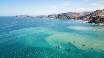 Vista aérea de pequeñas embarcaciones en el mar cerca de una costa desértica con cerros y variaciones en la tonalidad del agua