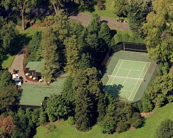 Vista aérea de una cancha de tenis verde con líneas blancas y red, rodeada de densa vegetación y árboles altos, junto a edificios y senderos