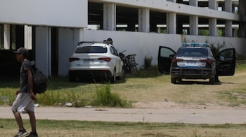 Un hombre camina a la izquierda, frente a un edificio blanco inconcluso. Dos automóviles, uno blanco y uno policial con puertas abiertas, están estacionados al lado
