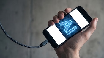 Una mano adulta sujeta un smartphone con la pantalla encendida mostrando un símbolo de inteligencia artificial azul. Un cable grueso conecta el teléfono.