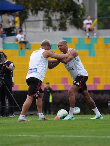 Dos hombres con camiseta blanca sin mangas y pantalón corto negro se sostienen por los hombros en una cancha de fútbol junto a un balón
