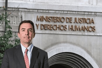 Un hombre de cabello oscuro y traje oscuro con corbata roja sonríe ligeramente frente al edificio del Ministerio de Justicia y Derechos Humanos, con follaje verde a la izquierda