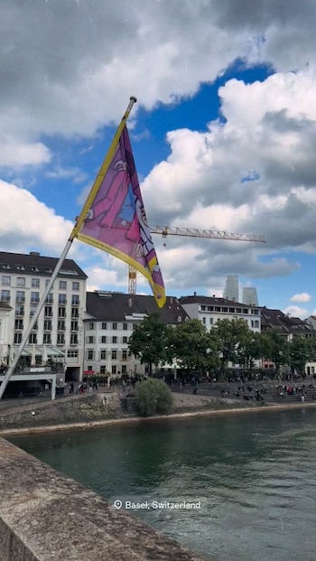 Vista aérea de un mástil con una bandera multicolor sobre un río en Basilea, Suiza, con edificios, árboles, personas y una grúa bajo un cielo nublado