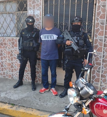 Dos policías armados con equipo táctico flanquean a un hombre detenido frente a una puerta metálica, con una motocicleta roja parcialmente visible