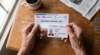 Dos manos arrugadas sostienen una tarjeta de descuento para autobús y una credencial INAPAM. En la mesa hay una taza de café, un periódico y monedas.