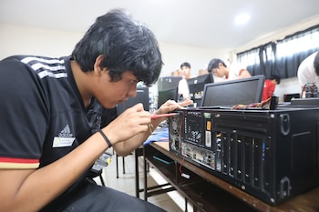 Un joven de cabello oscuro y camiseta negra Adidas, concentrado, usa un destornillador para trabajar en el hardware trasero de una torre de computadora en un aula con otros estudiantes