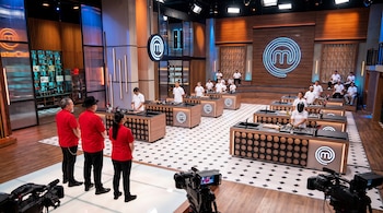 Un estudio de televisión con estaciones de cocina, personas vestidas de chef, tres jueces en camisa roja y dos cámaras de grabación en primer plano.